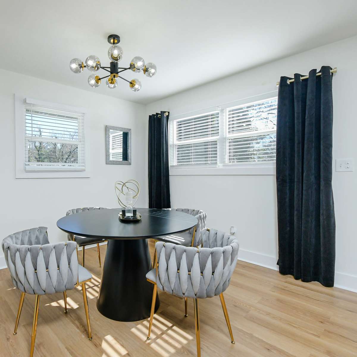 A modern dining space featuring a round black table with gold accents and four tufted gray chairs, illuminated by a decorative pendant light.