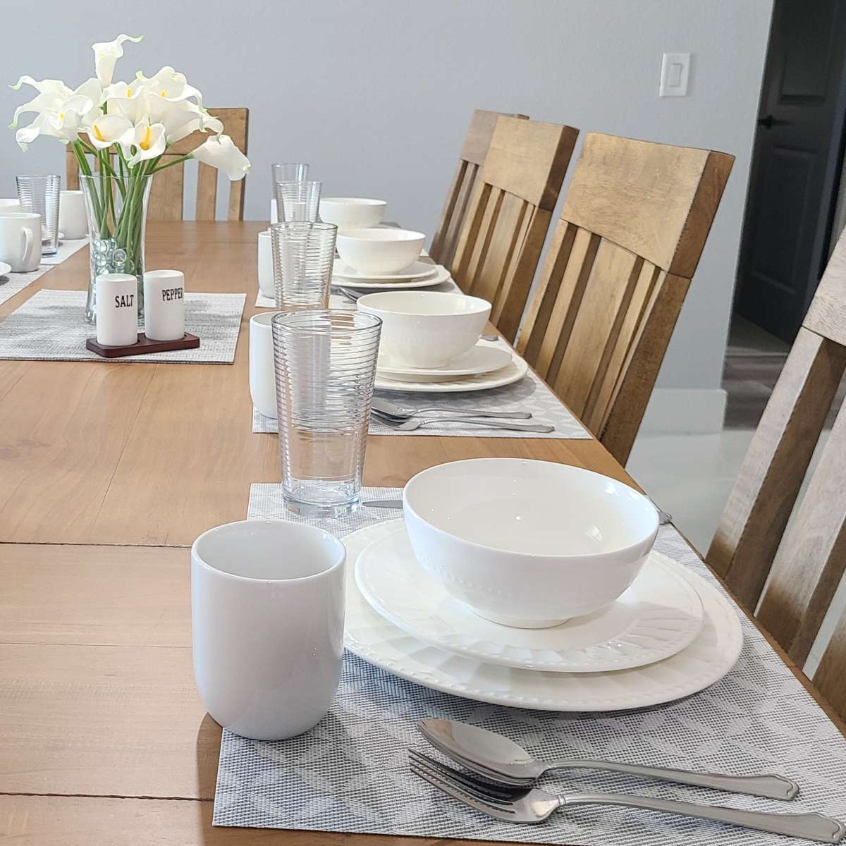 A dining table is set for a meal with plates, bowls, glasses, and a vase of flowers arranged down the center.