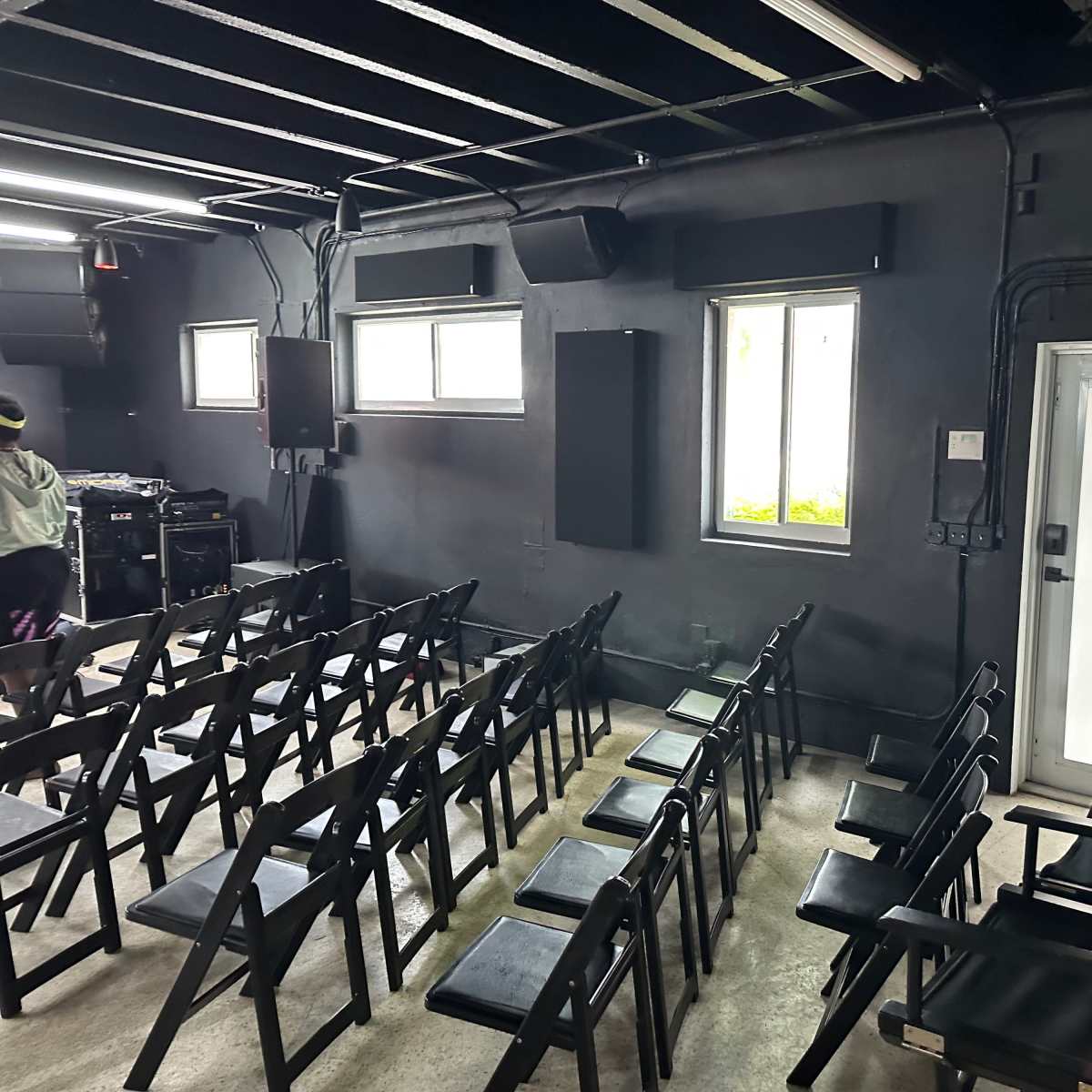 A dimly lit room set up with rows of black folding chairs facing a wall of speakers and a door.