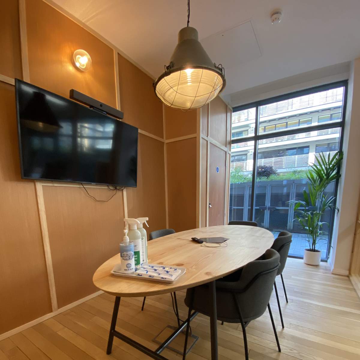 A modern meeting room features a wooden table with chairs, a wall-mounted TV, and a window overlooking greenery.