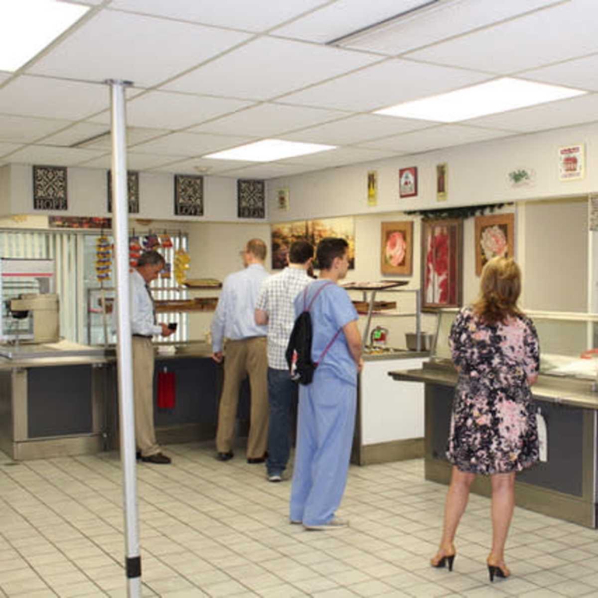 A group of people stands in line at a cafeteria with tables and food displays visible in the background.