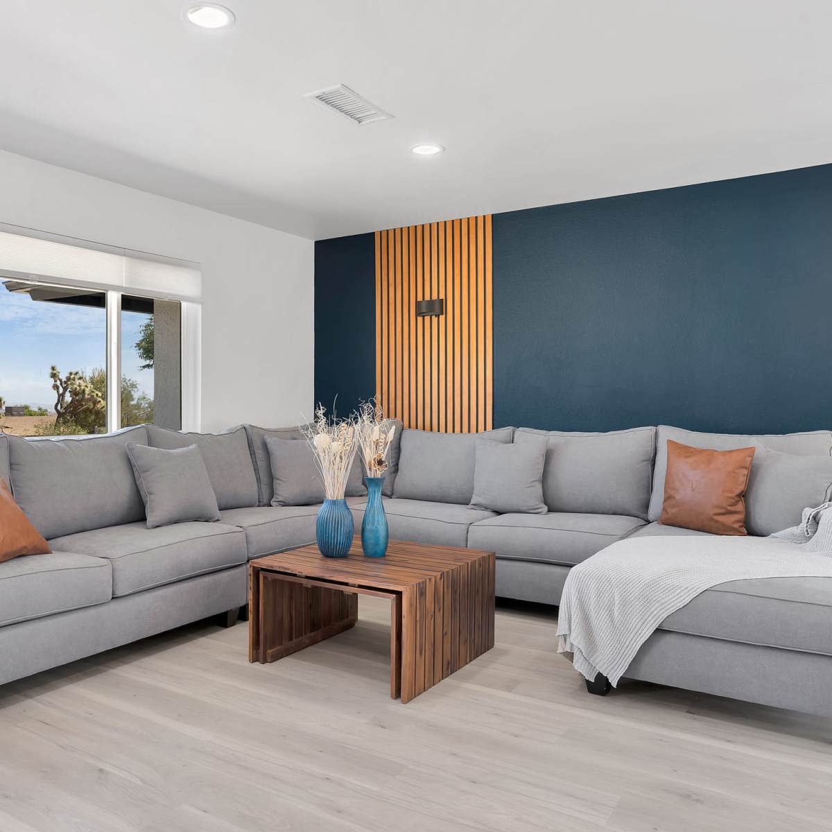 A modern living room featuring a gray sectional sofa, a wooden coffee table, and a textured dark blue accent wall, with natural light coming through a large window.