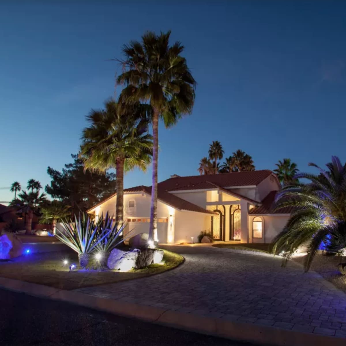 A modern house with a tiled roof is surrounded by palm trees and illuminated by landscape lighting at dusk.