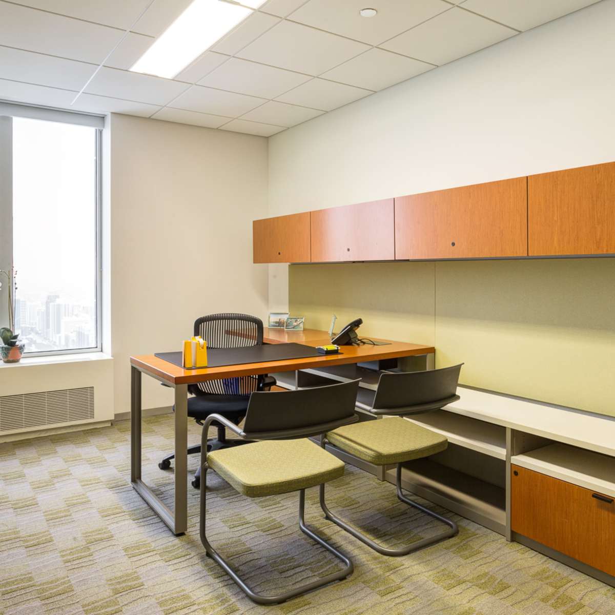 A modern office featuring a wooden desk with a chair and two additional chairs, a wall-mounted storage unit, and a large window offering a view of the city.