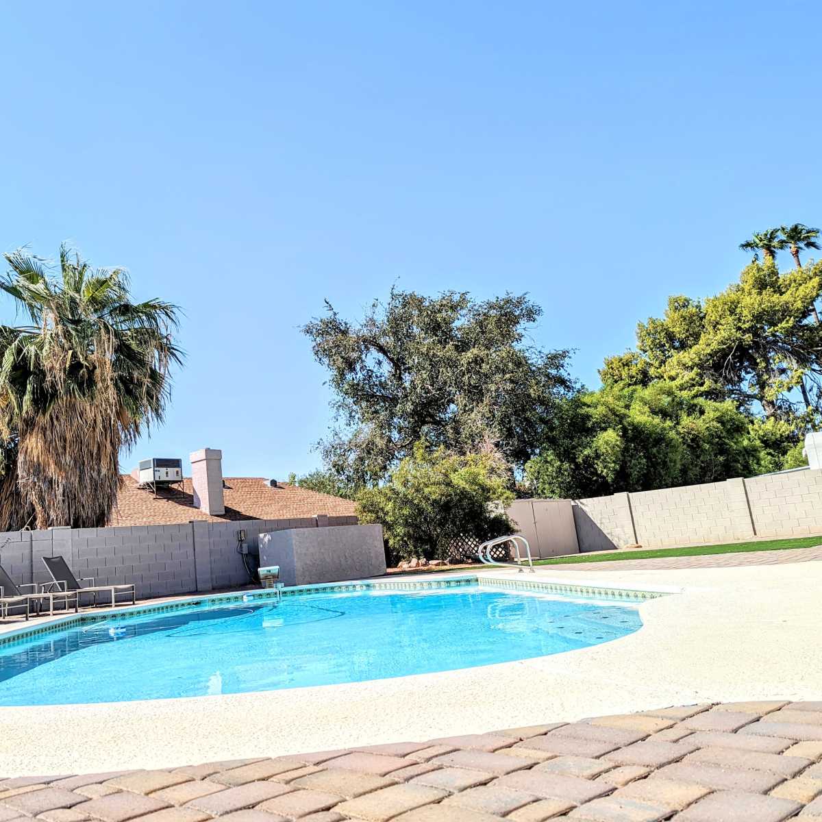 A clear swimming pool surrounded by a patio and several lounge chairs, with palm trees and other greenery in the background.