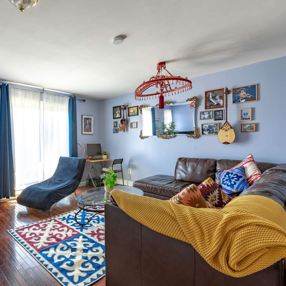 A cozy living room features a dark leather sectional sofa, a blue chaise lounge, a large flat-screen TV, and a patterned area rug, with walls adorned with various framed pictures.