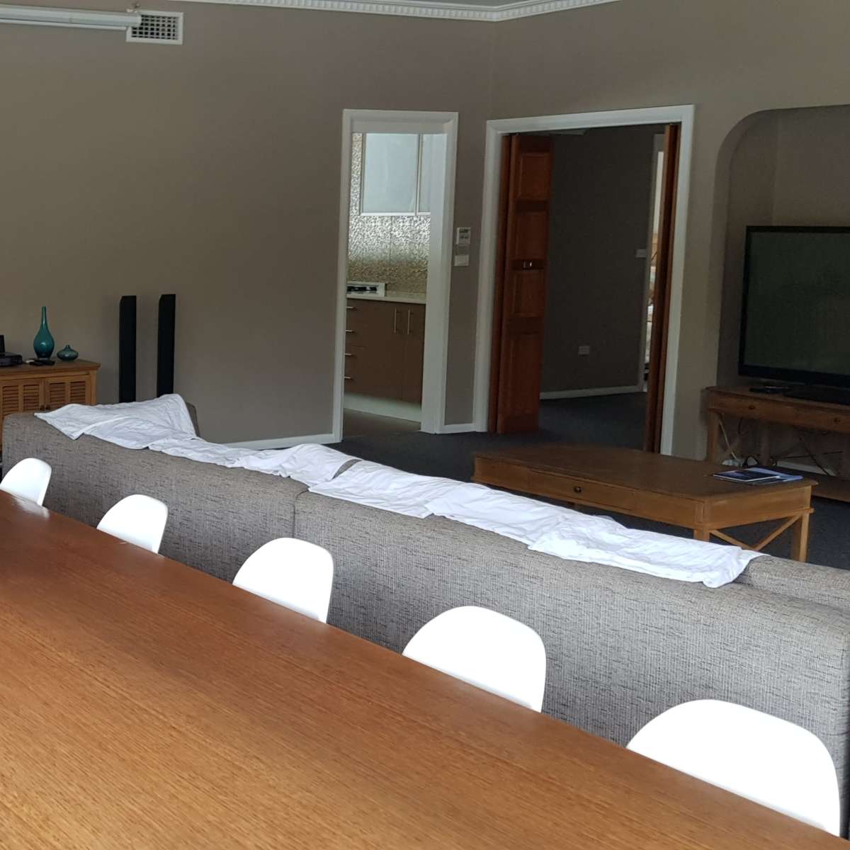 A spacious living area with a long wooden dining table, white chairs, a gray sofa, and a coffee table, all set in a room with light walls and dark carpet.