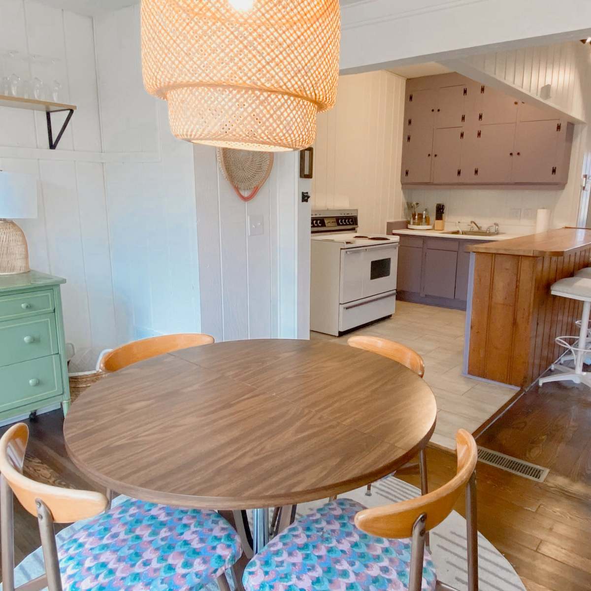 A round wooden dining table with pink and blue patterned chairs is placed in a bright kitchen area featuring a green sideboard and a large pendant light above.
