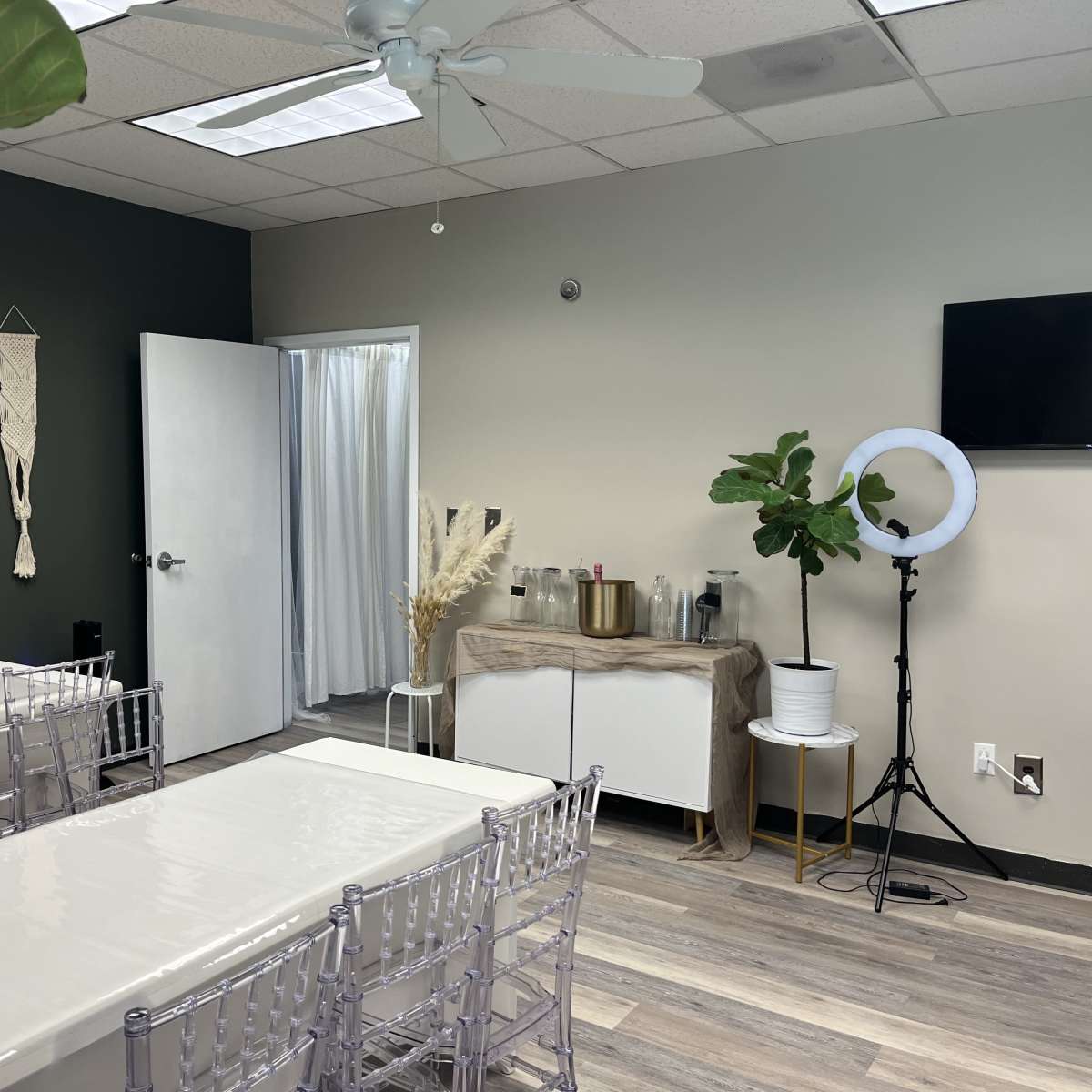A neatly arranged meeting room with a large table, transparent chairs, a plant, a wall-mounted TV, and a bar area featuring drinks and decorative elements.