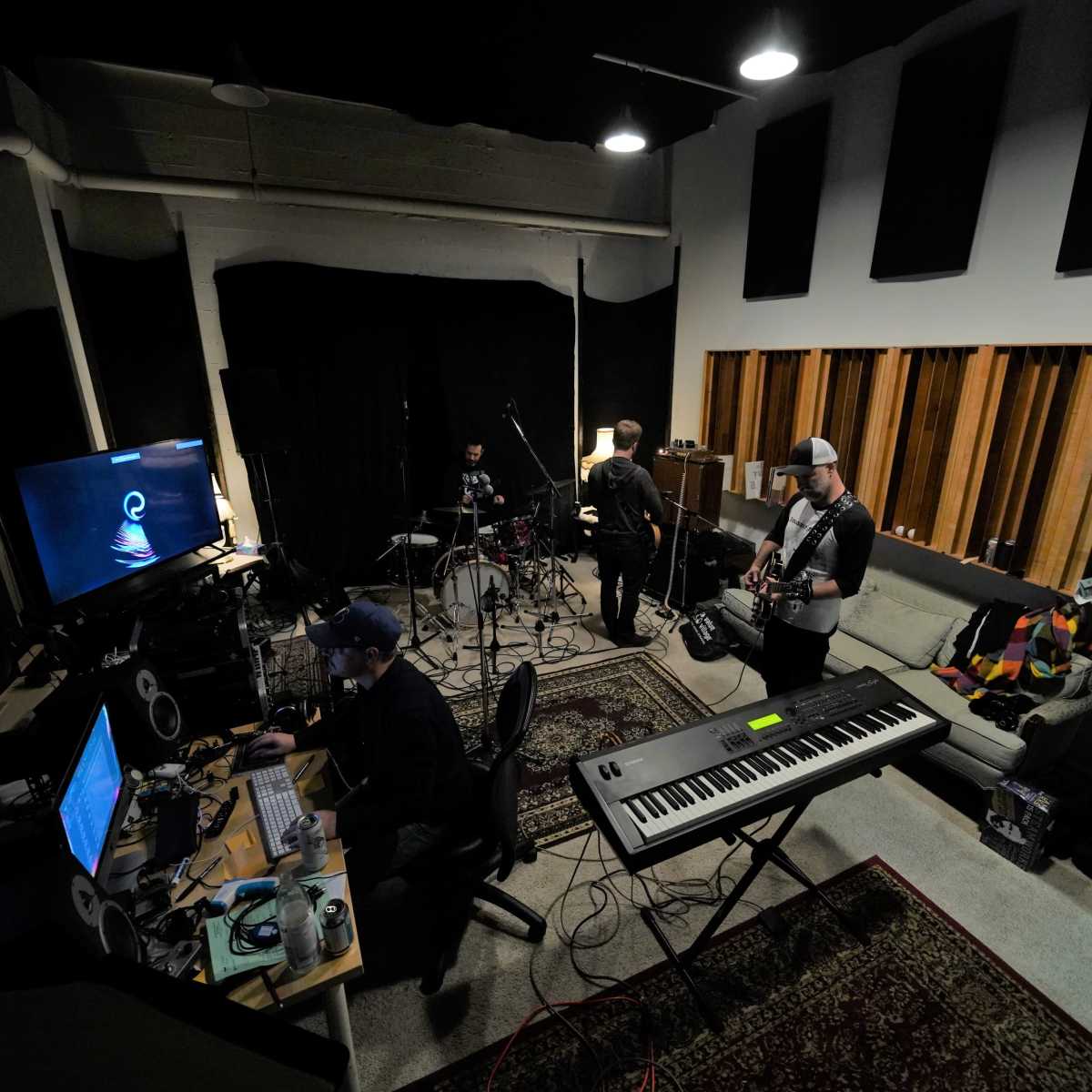 A group of musicians collaborates in a studio, with one on the keyboard, another on drums, and a guitarist in the background, surrounded by various audio equipment.