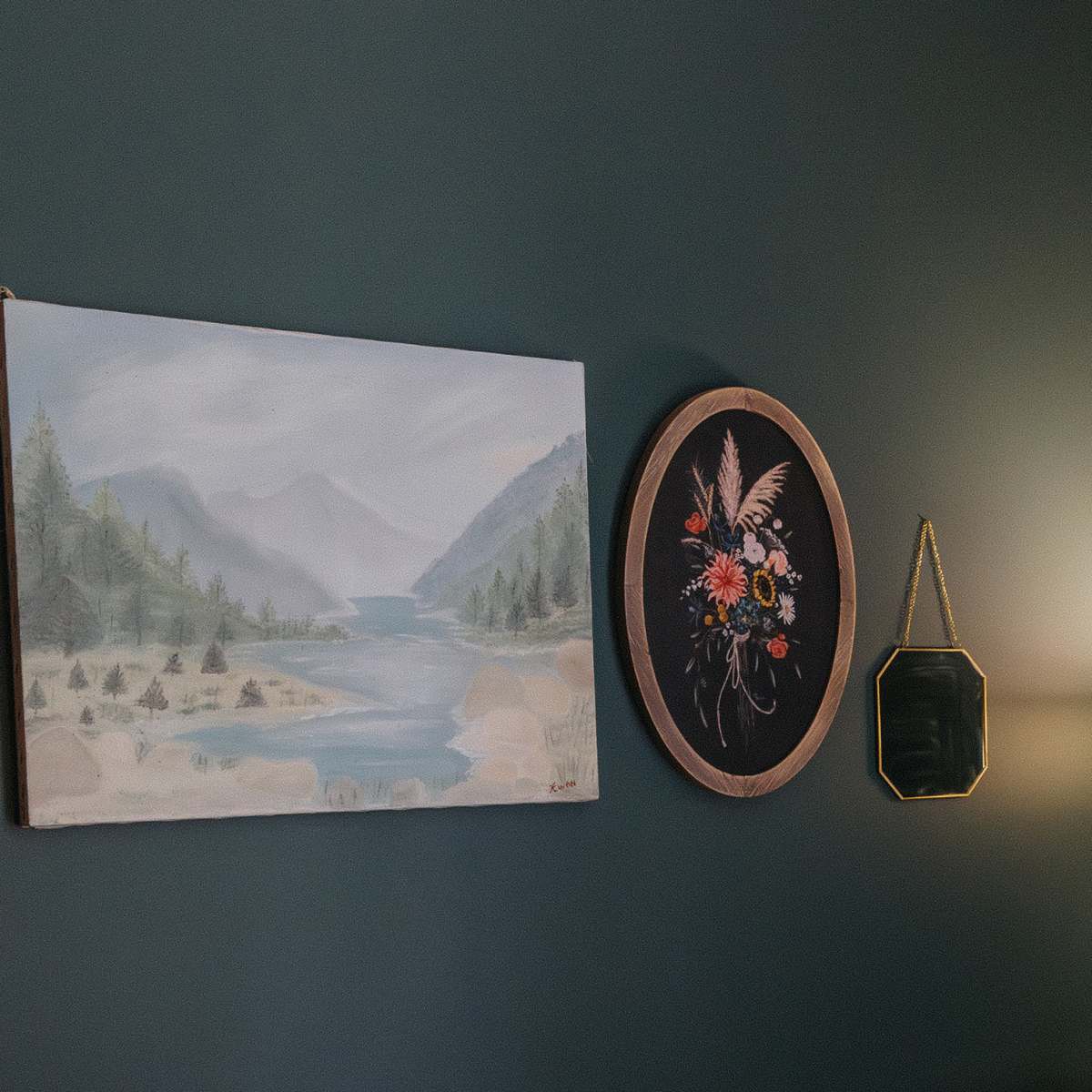 A wall adorned with a landscape painting of a river and mountains, framed artwork of flowers, and decorative mirrors.