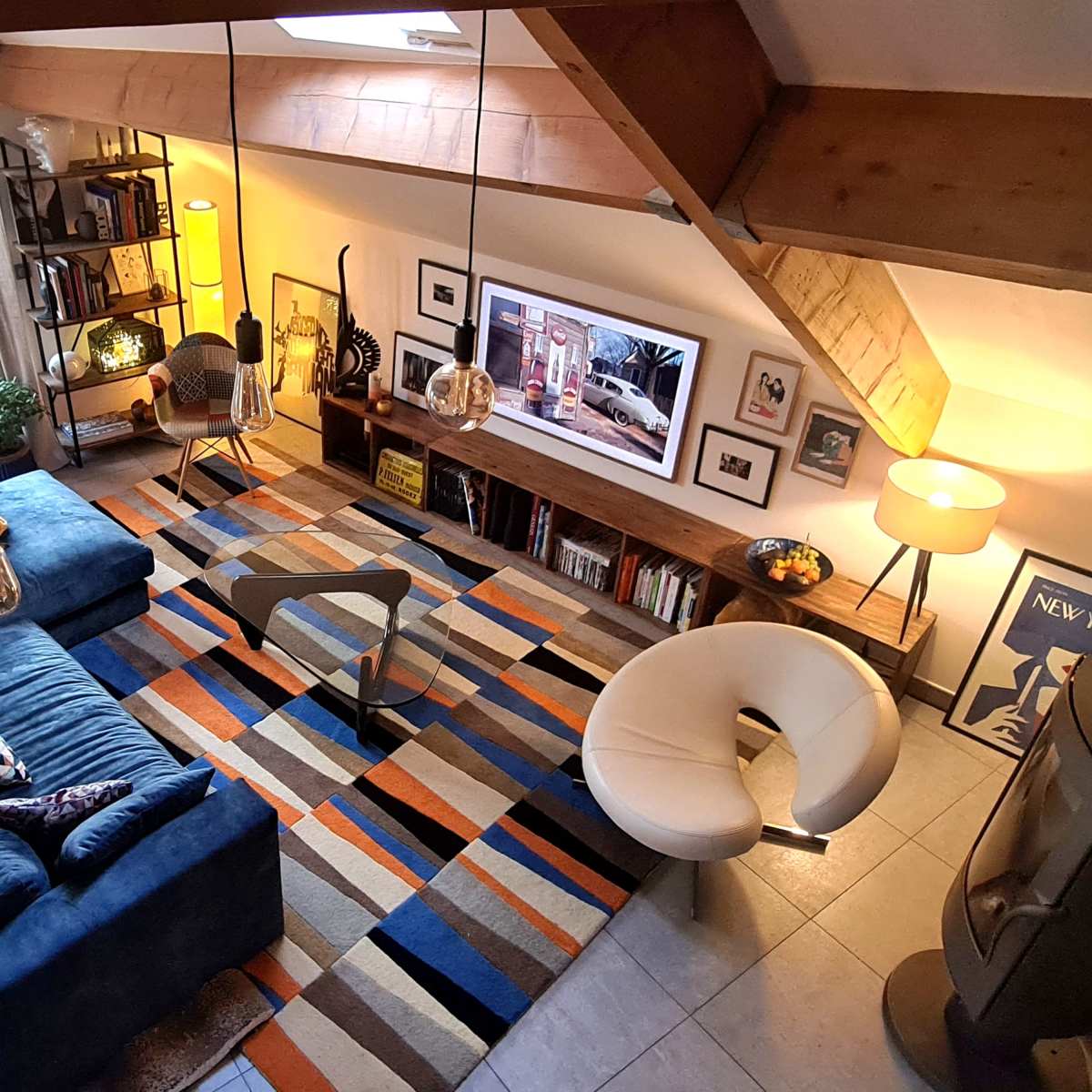 A stylish living room featuring a blue sectional sofa, a round white chair, a patterned area rug, and a wall-mounted television above a wooden shelf, with large windows allowing natural light to enter.