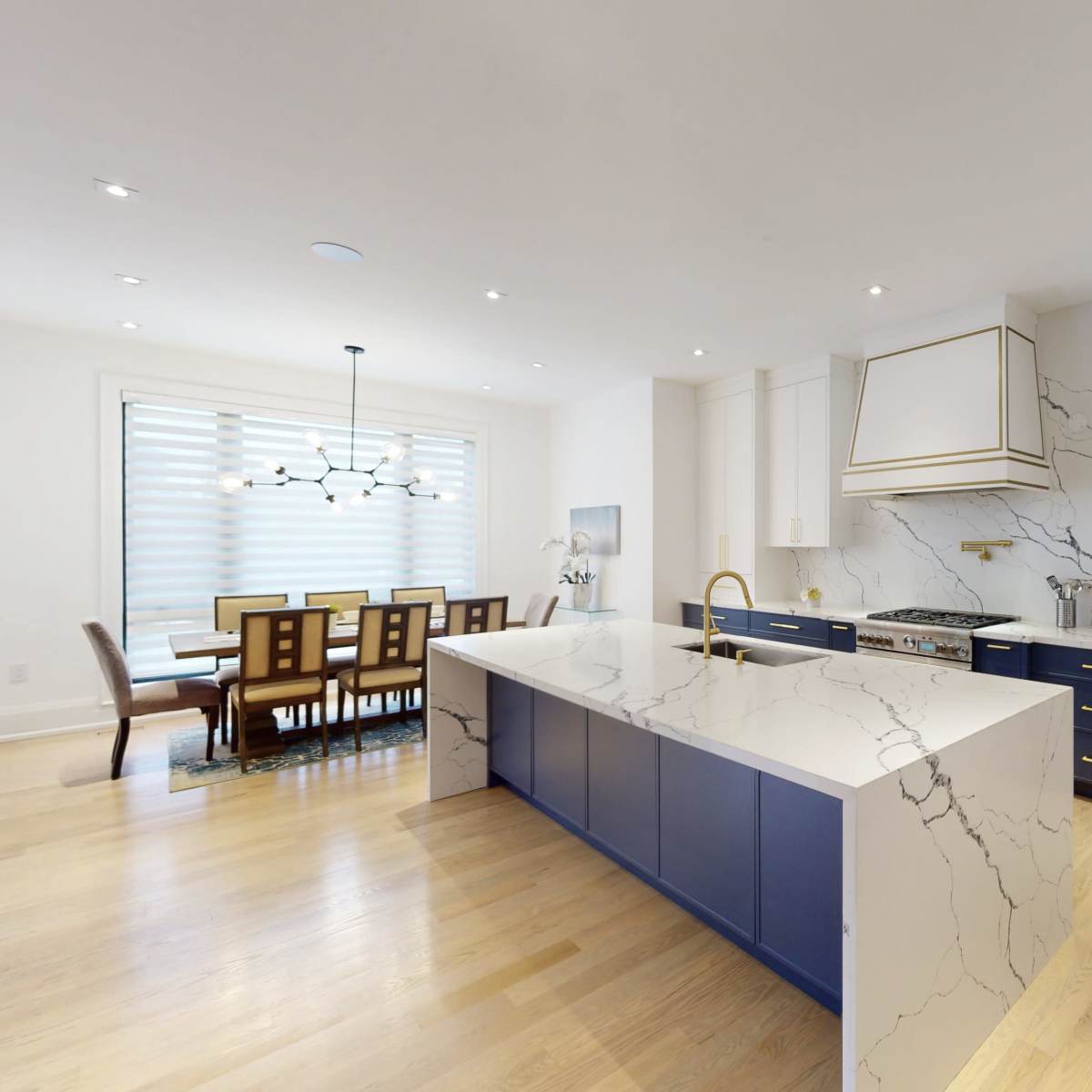 A modern kitchen features a large island with a white and blue marble countertop, surrounded by white cabinetry and gold accents, with a dining area visible in the background.