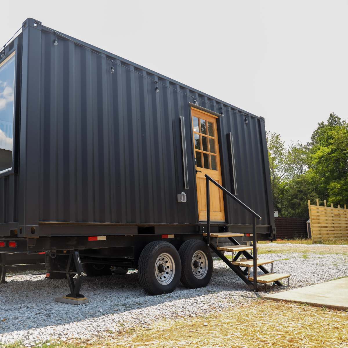 A black shipping container converted into a mobile tiny house is positioned on a gravel surface with a wooden staircase leading to its door.