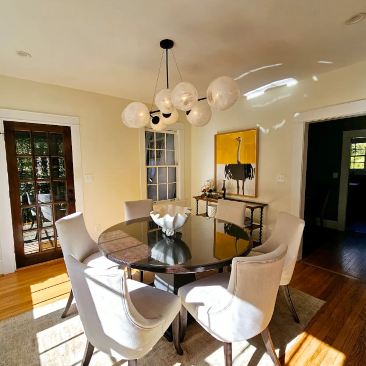 A round table with six upholstered chairs is centered in a well-lit dining room featuring a chandelier and artwork on the walls.