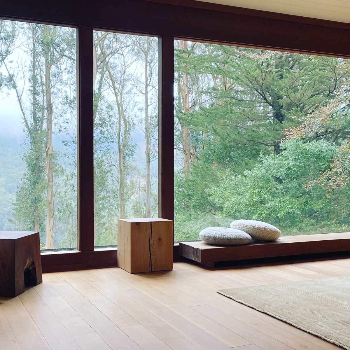A spacious room with large windows overlooking a forested landscape, featuring a chair with a pillow and a wooden bench.