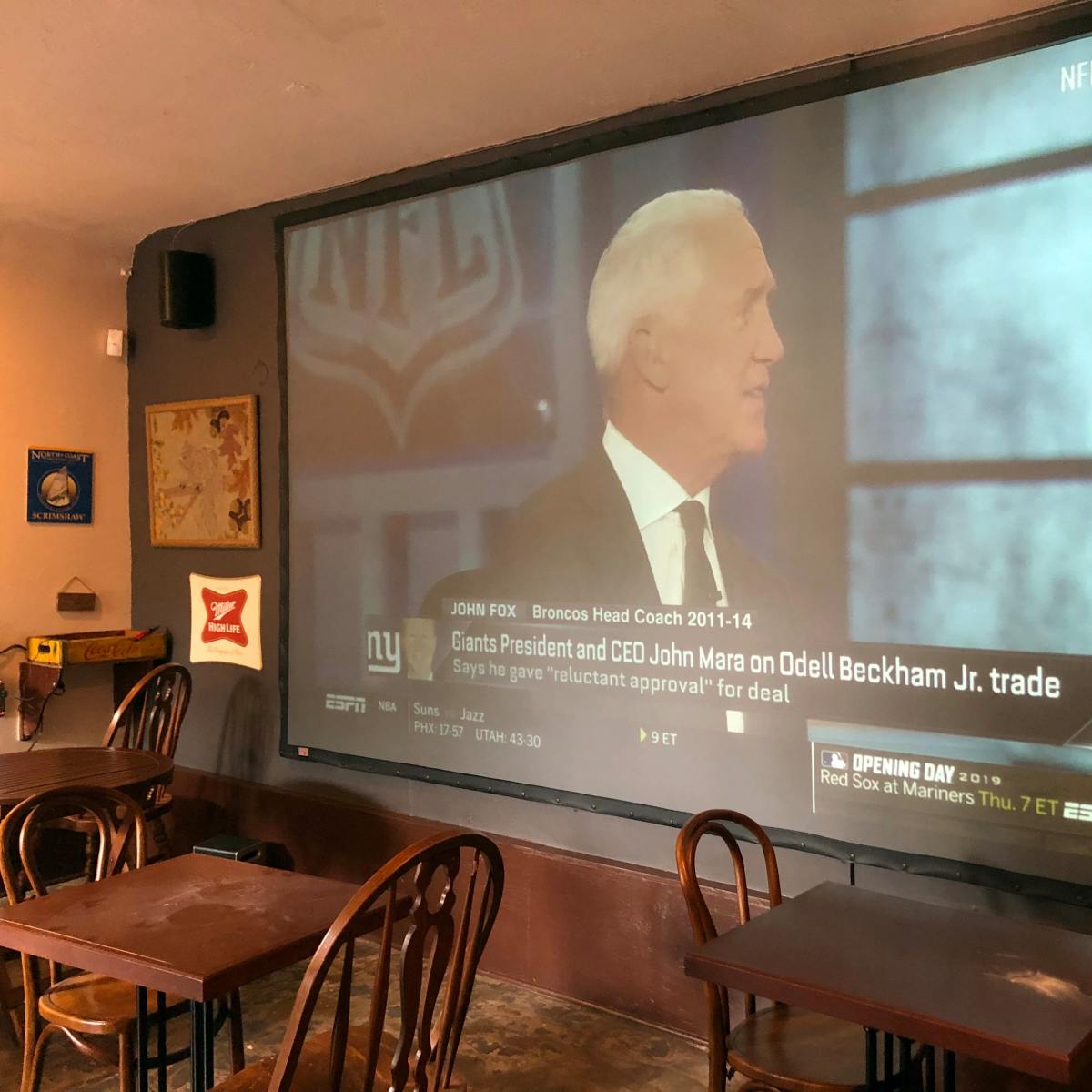 A large screen displays a sports news segment featuring John Fox discussing NFL trades in a bar setting with wooden tables and chairs.