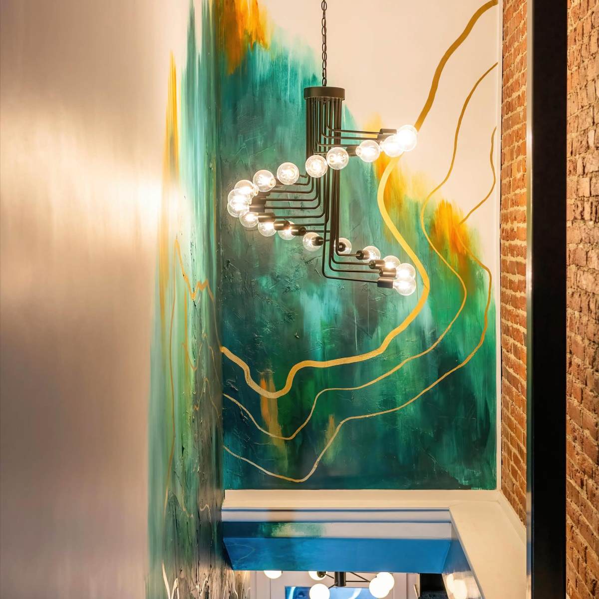 A colorful mural with green and gold accents adorns the wall of a well-lit stairway featuring a modern chandelier.