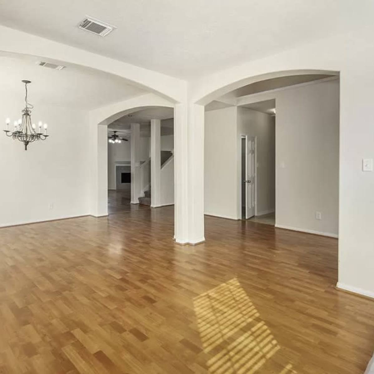 A spacious room with hardwood flooring, archway openings, and a chandelier, leading to a hallway and another area in the background.