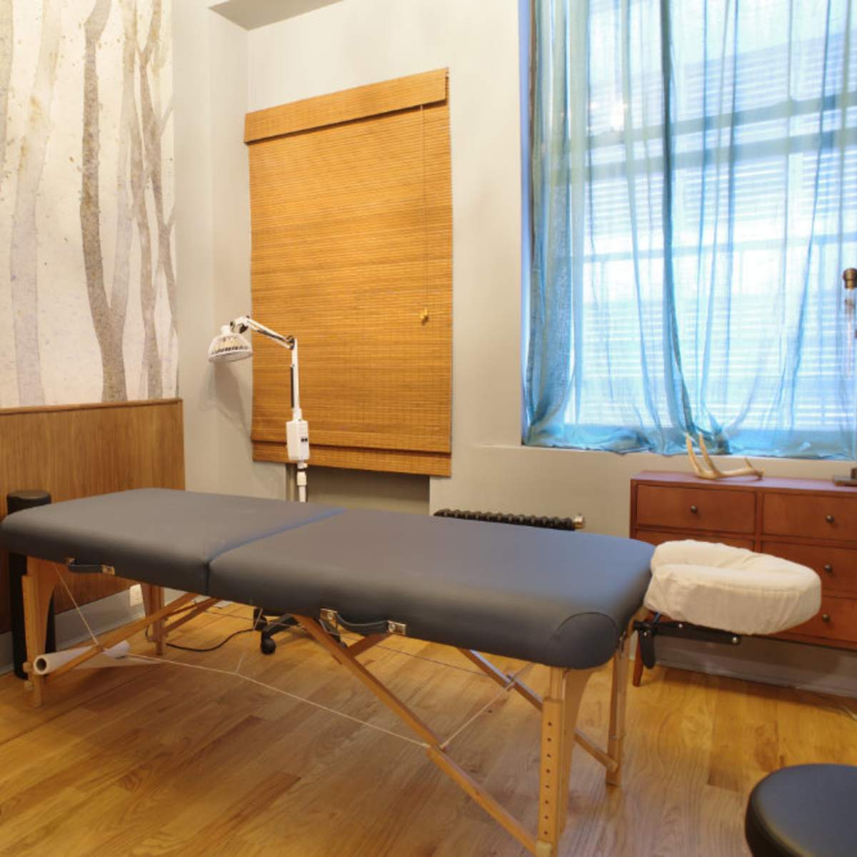 A massage therapy room features a wooden table, a chair, and a large window with sheer curtains.