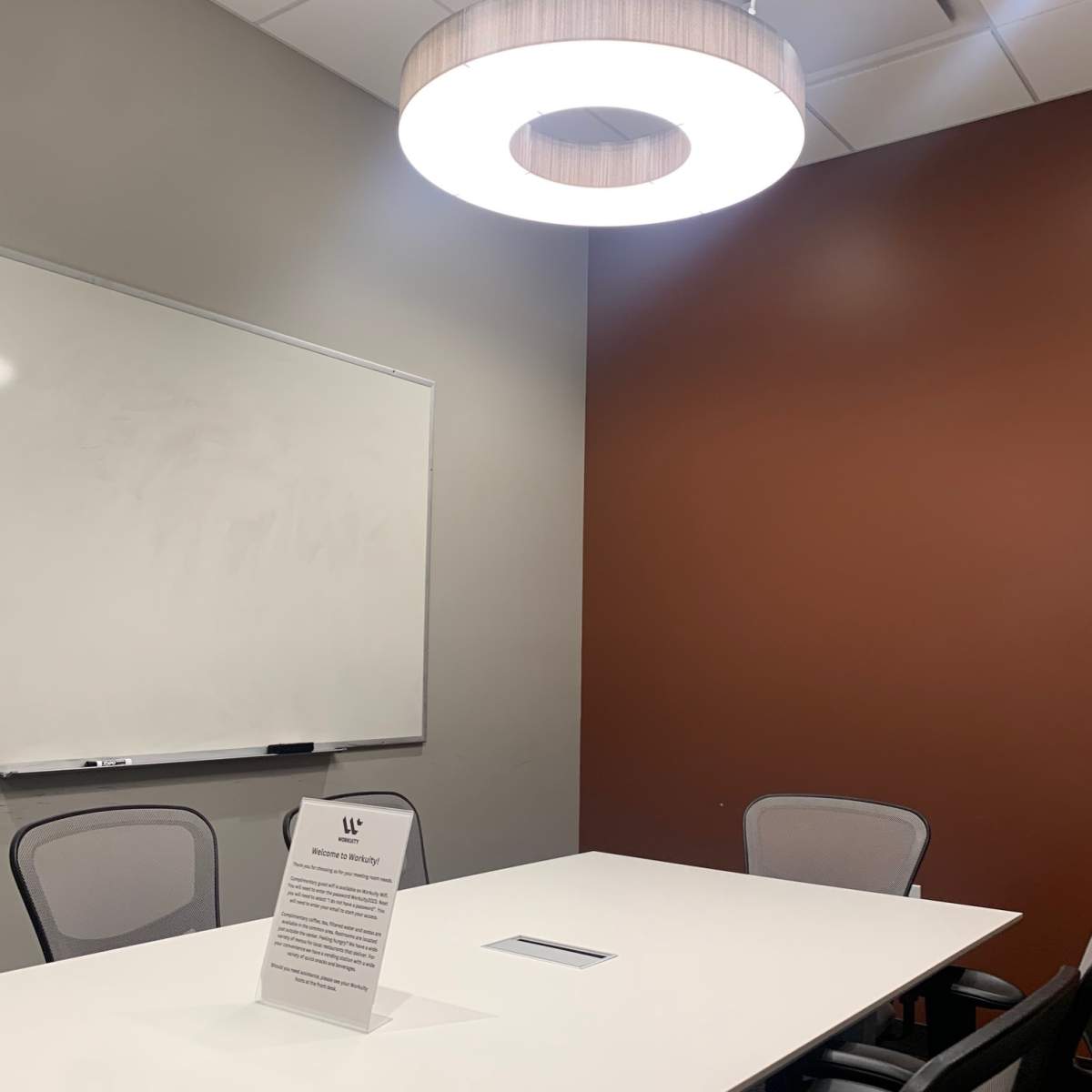 A meeting room features a whiteboard, a contemporary light fixture, a rectangular table, and four chairs, with a sign displayed on the table.