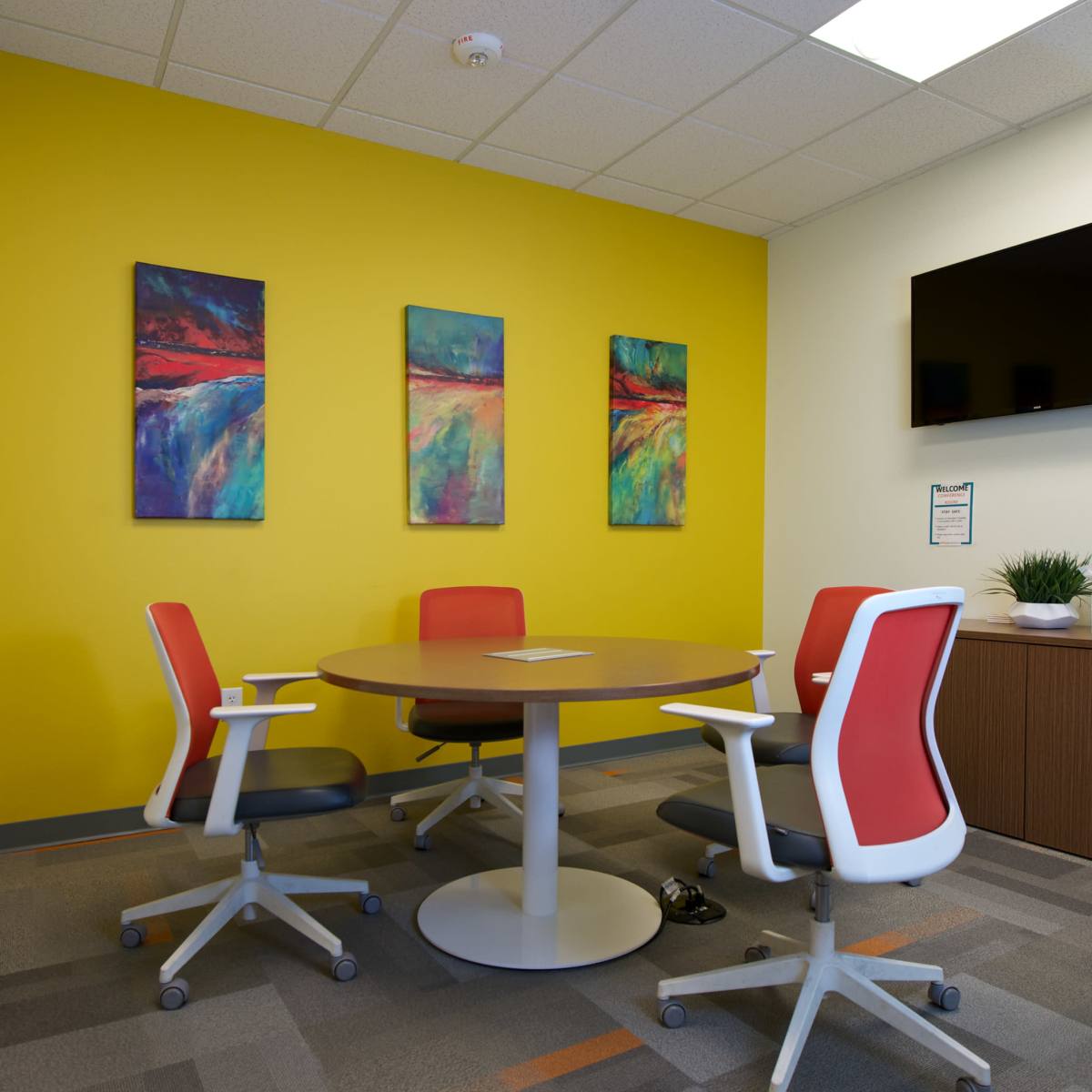 A meeting room features a round table surrounded by four orange and gray rolling chairs against a bright yellow wall adorned with three colorful paintings.