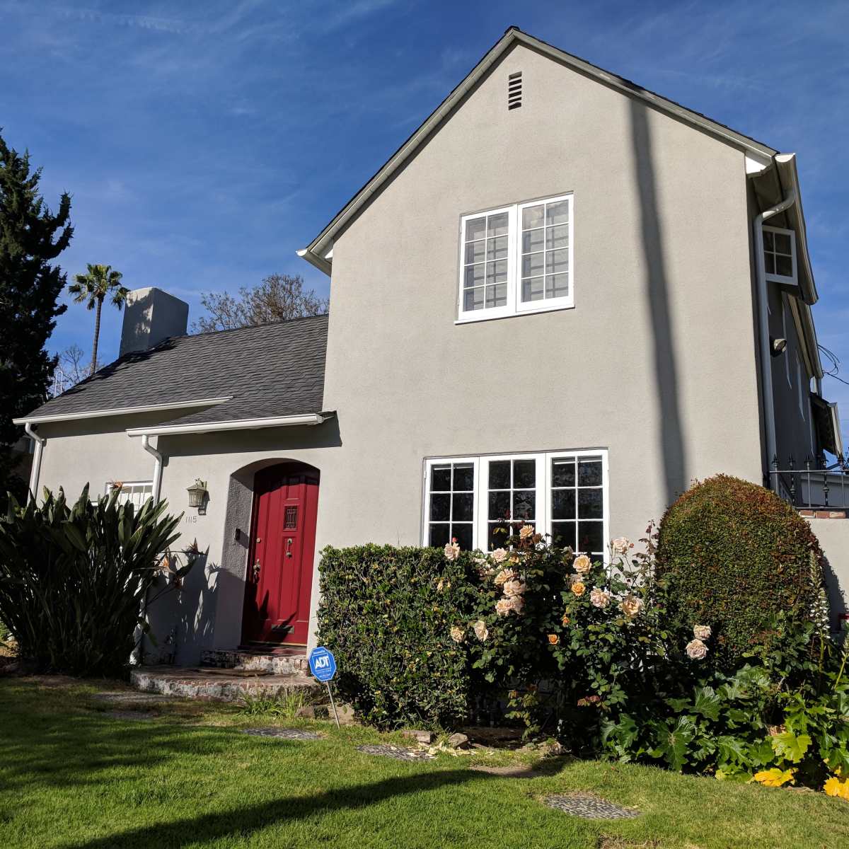 A two-story gray house with a red door is surrounded by greenery and flowers in the garden.