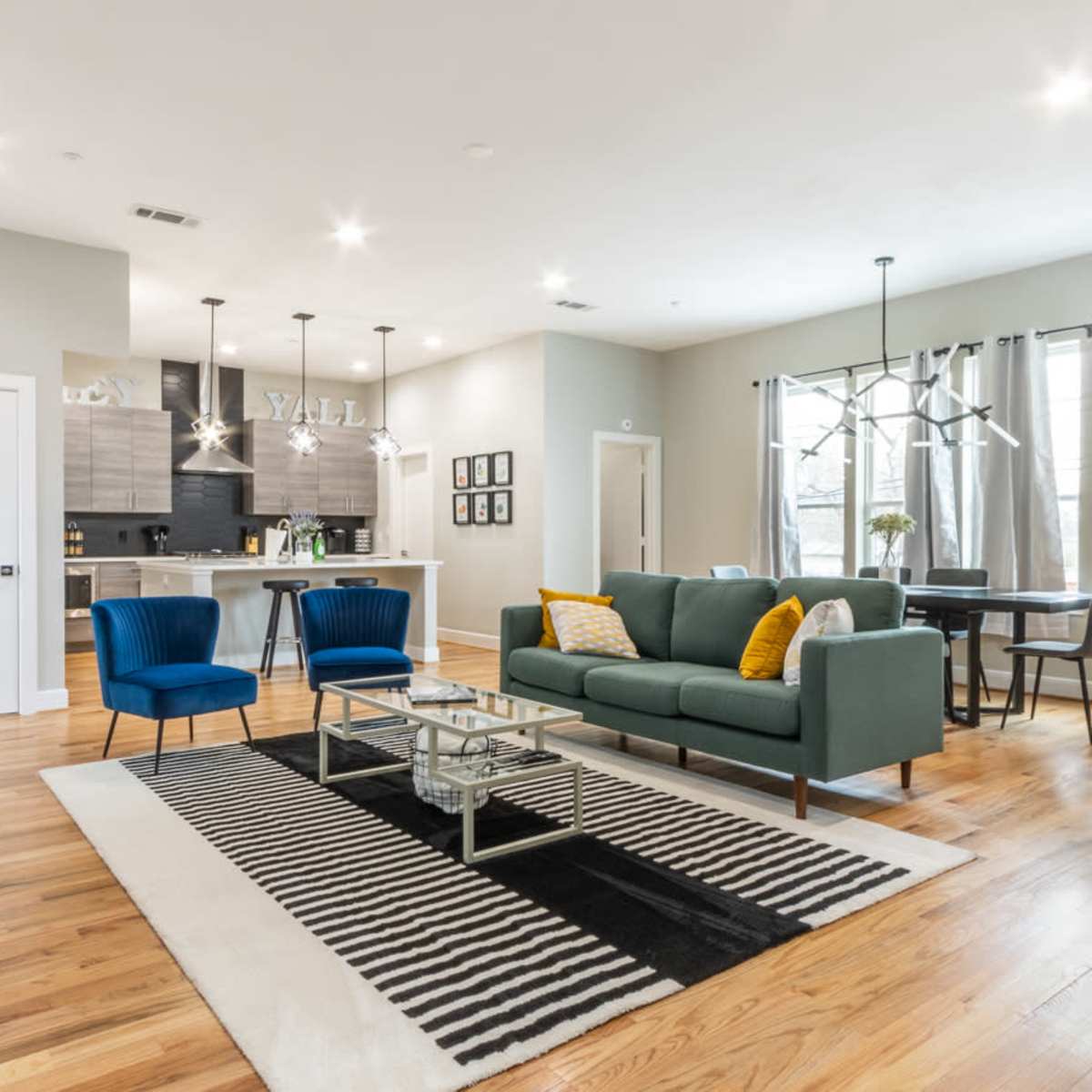 A modern living room with a green sofa, two blue chairs, a coffee table, and wooden flooring, along with an adjacent kitchen area visible in the background.