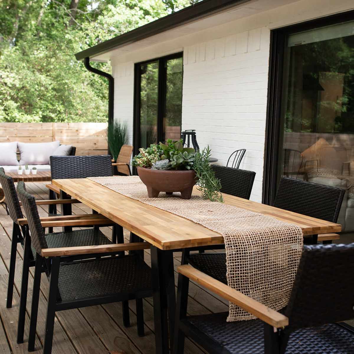 A wooden dining table with black chairs is set on a wooden deck, surrounded by greenery and adjacent to a modern home.
