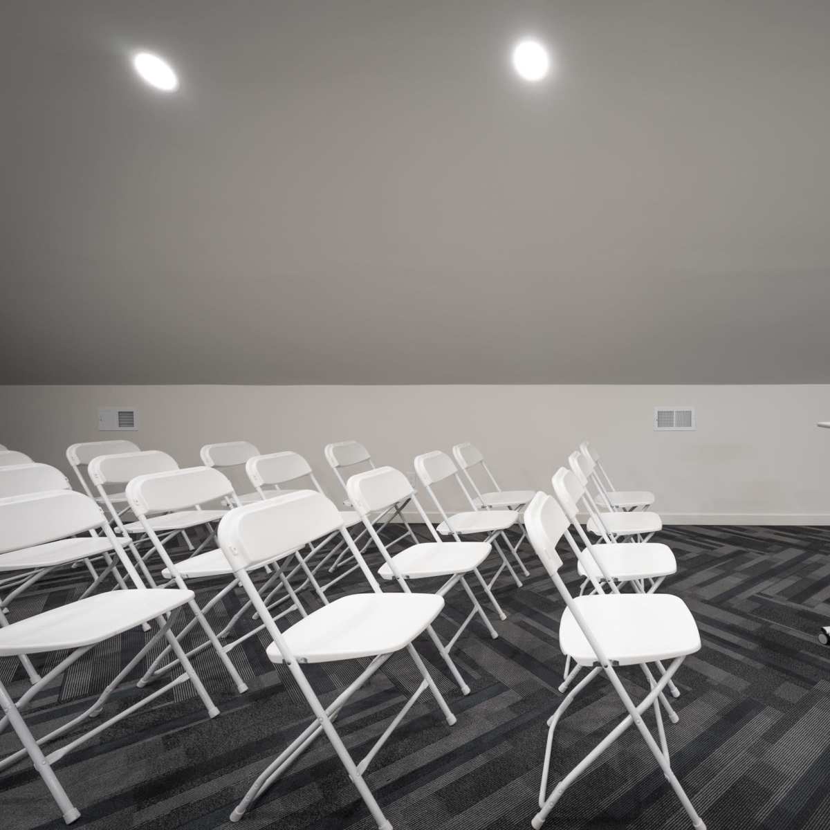 A small, minimalist meeting room features rows of white folding chairs and a podium with a stool on a patterned carpet.