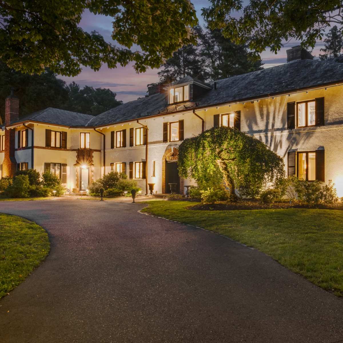 A large, two-story house with a landscaped front yard is illuminated at dusk, showcasing its windows and architectural details.