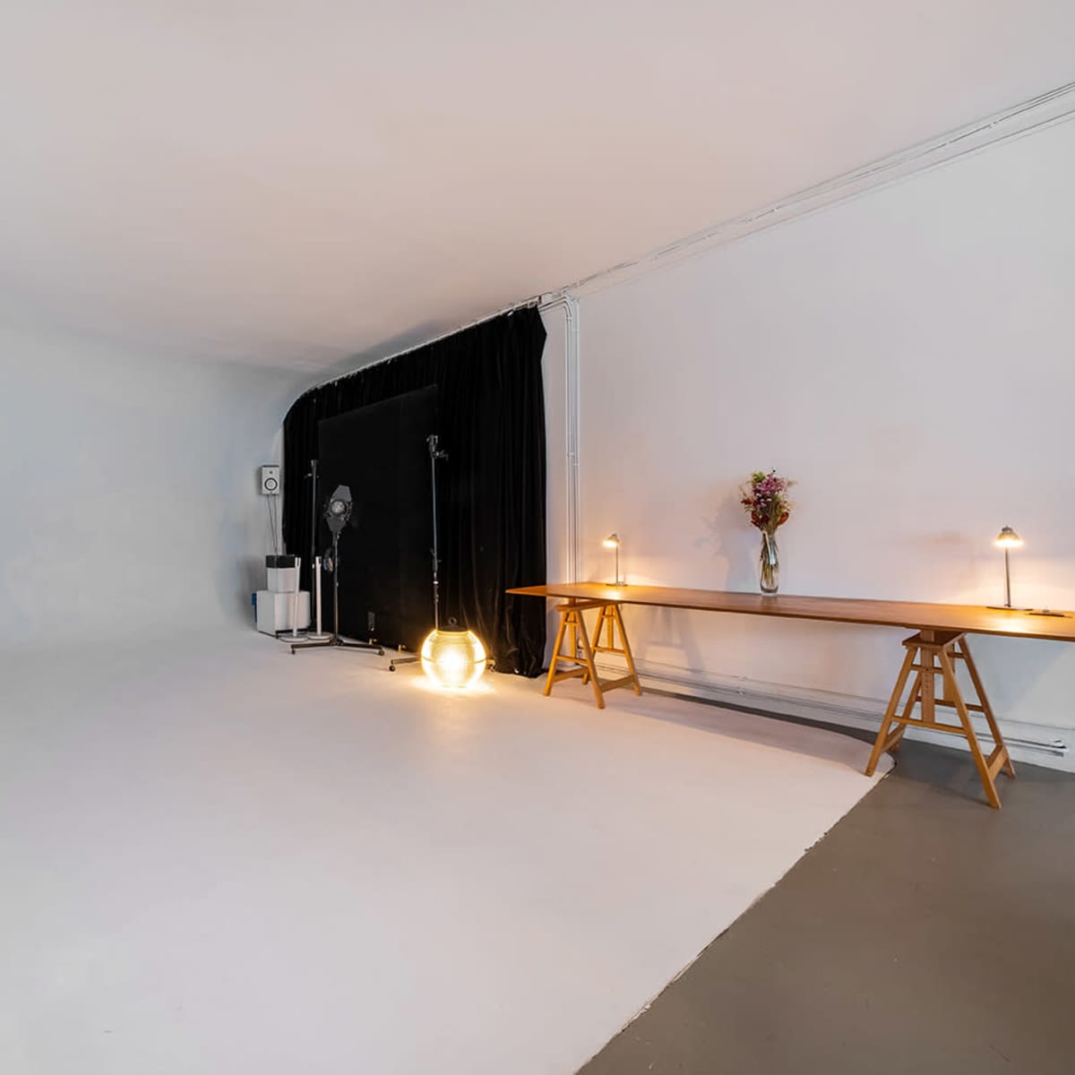 A spacious, minimalist studio with a plain white backdrop, black curtains on one side, and a wooden table with lamps and a vase of flowers.