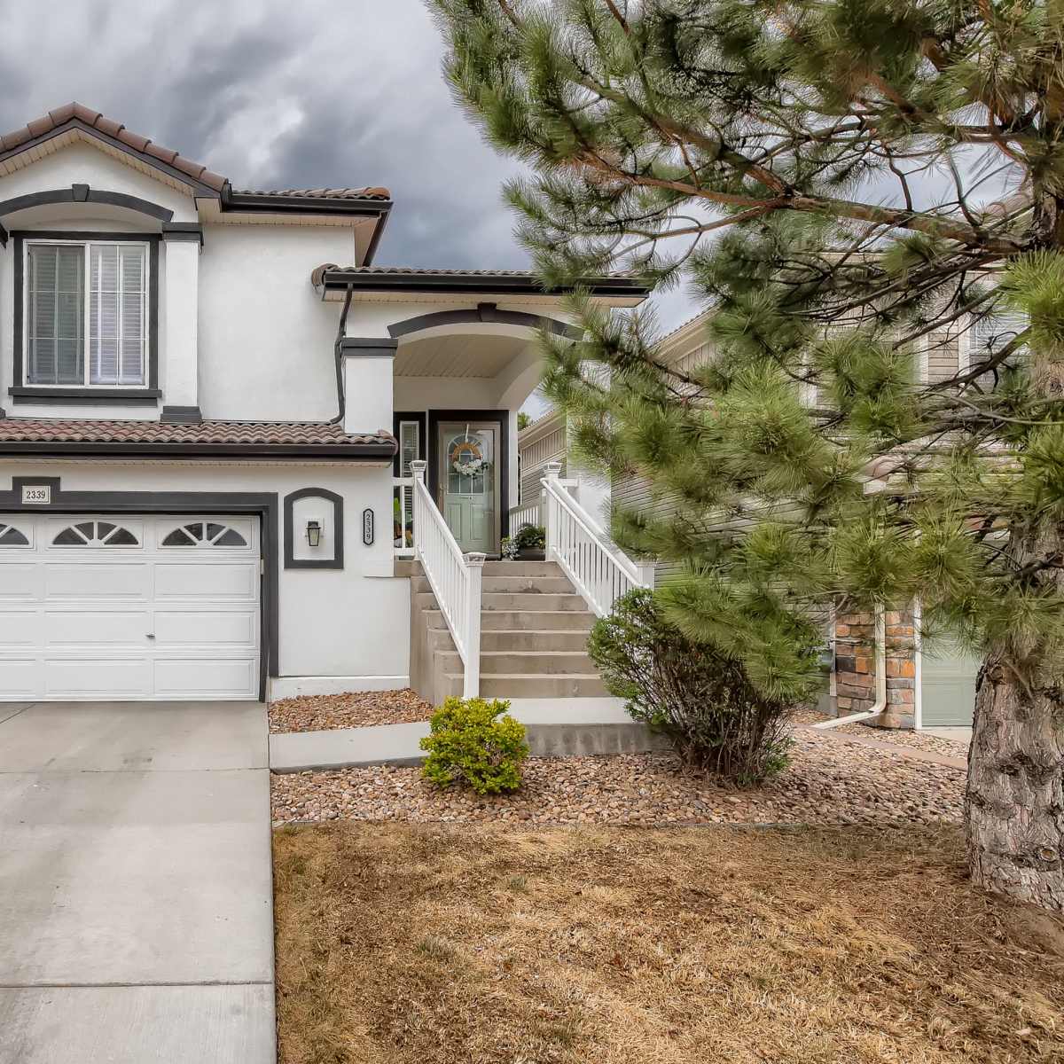 A two-story house with a white exterior and a tiled roof is situated on a paved driveway, flanked by neatly maintained landscaping.