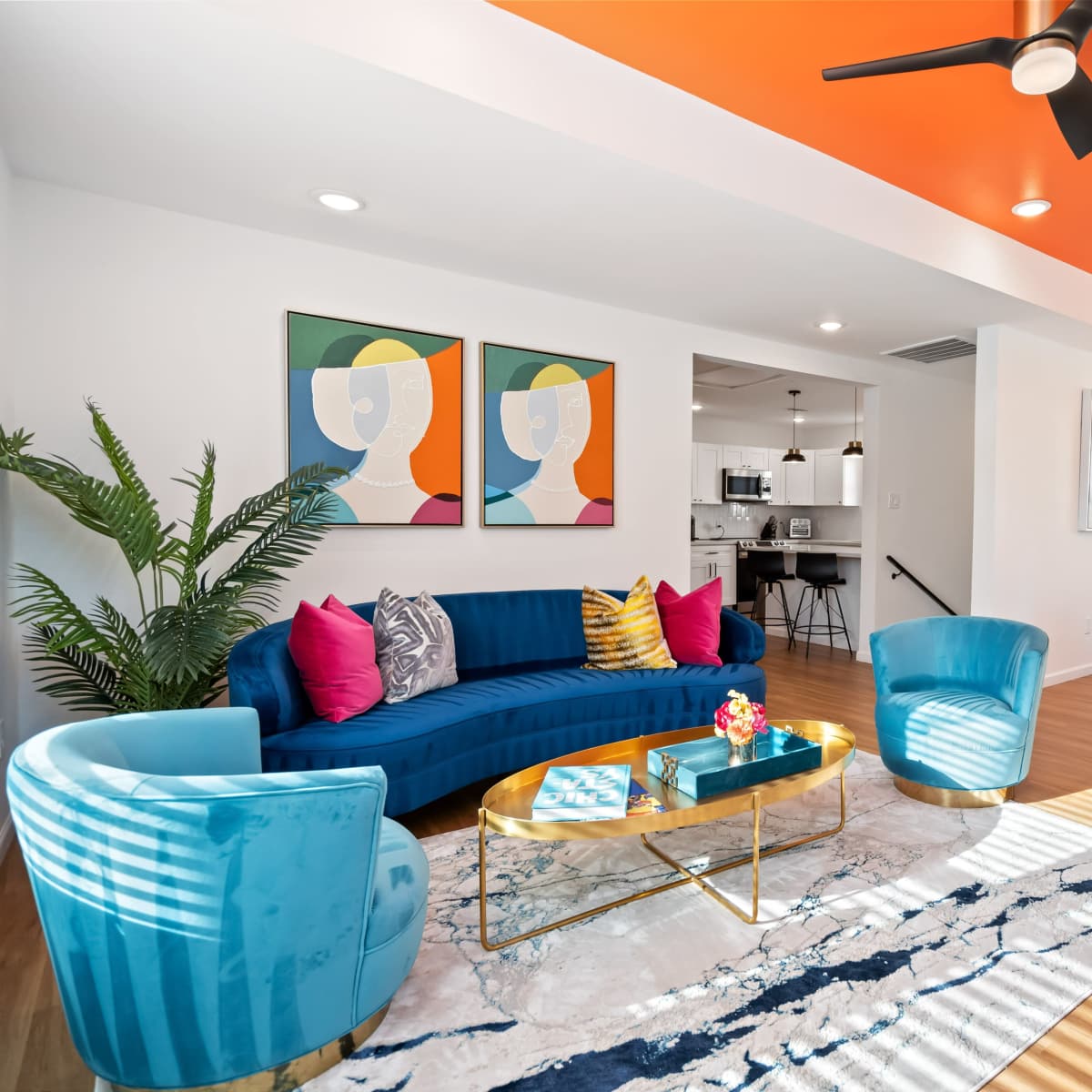 The living room features a blue sofa, two matching chairs, a circular gold coffee table, and colorful artwork on the walls, with a bright orange ceiling above.