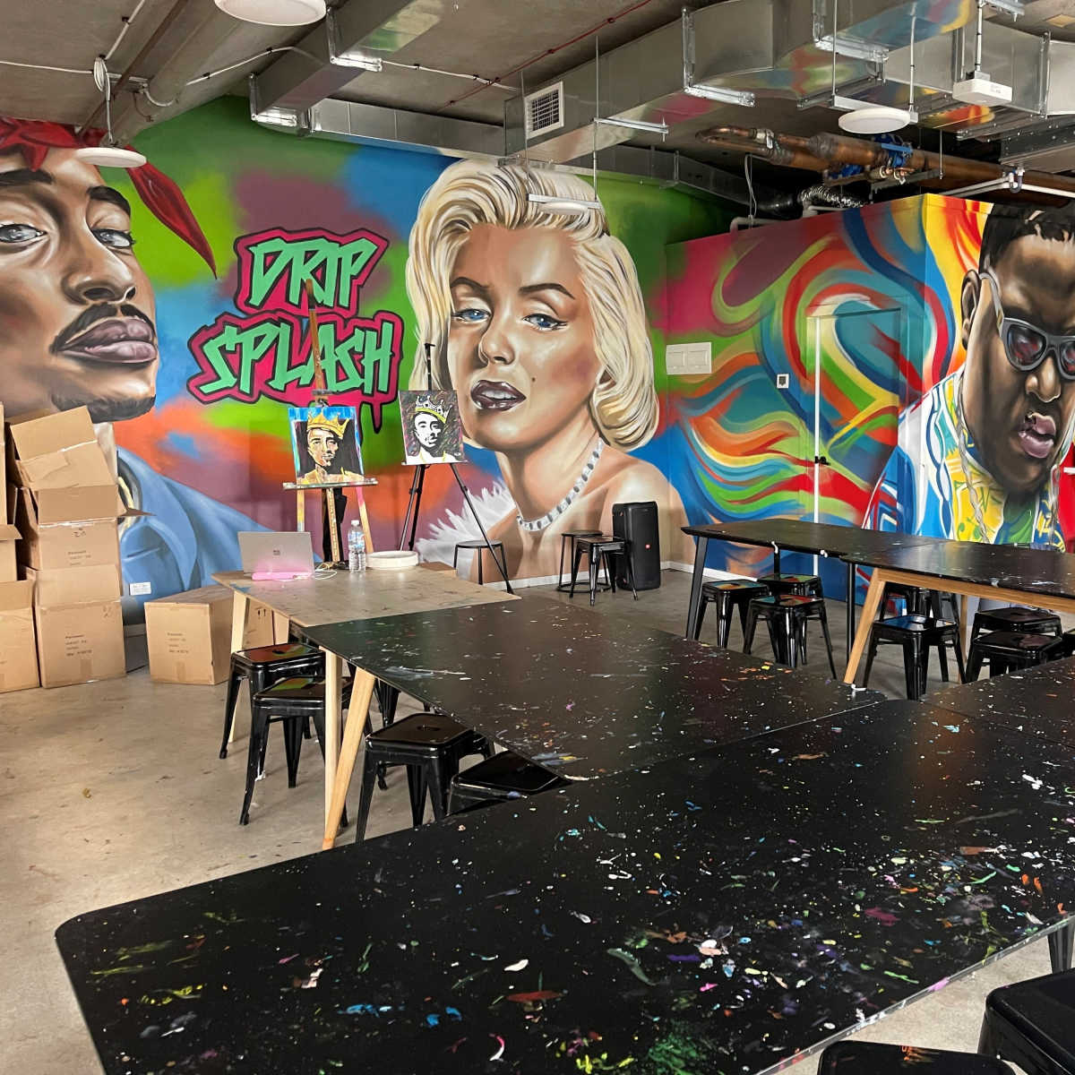 An art studio with colorful murals of iconic figures on the walls, surrounded by rows of black tables and chairs.
