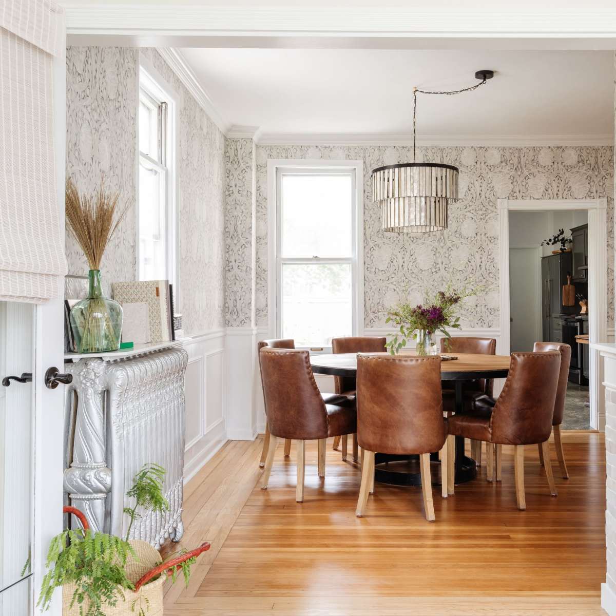 A dining room features a wooden table surrounded by brown leather chairs, with patterned wallpaper and a modern light fixture above.