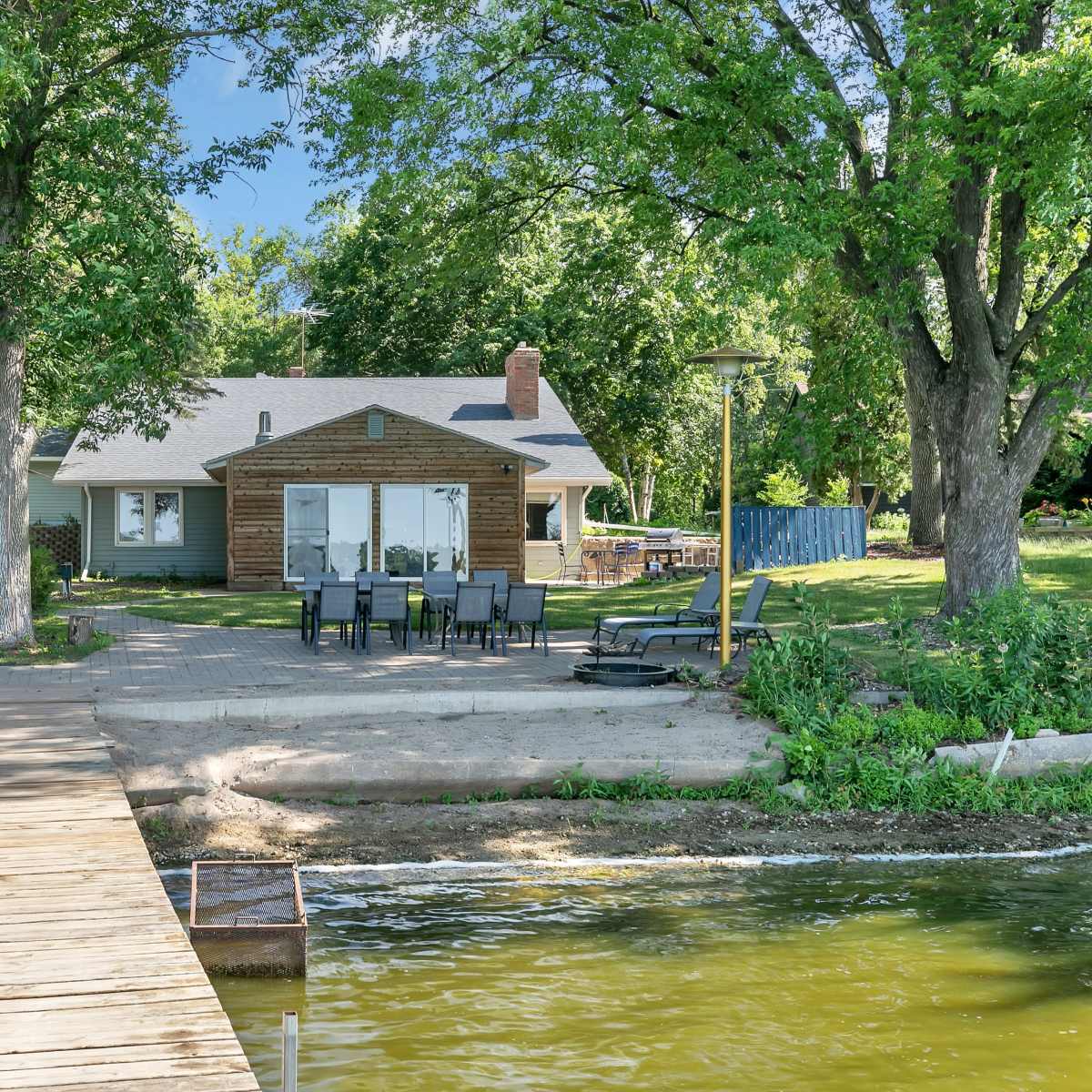 A wooden dock extends over the water toward a house set back from the shore, surrounded by trees and a patio area with outdoor seating.