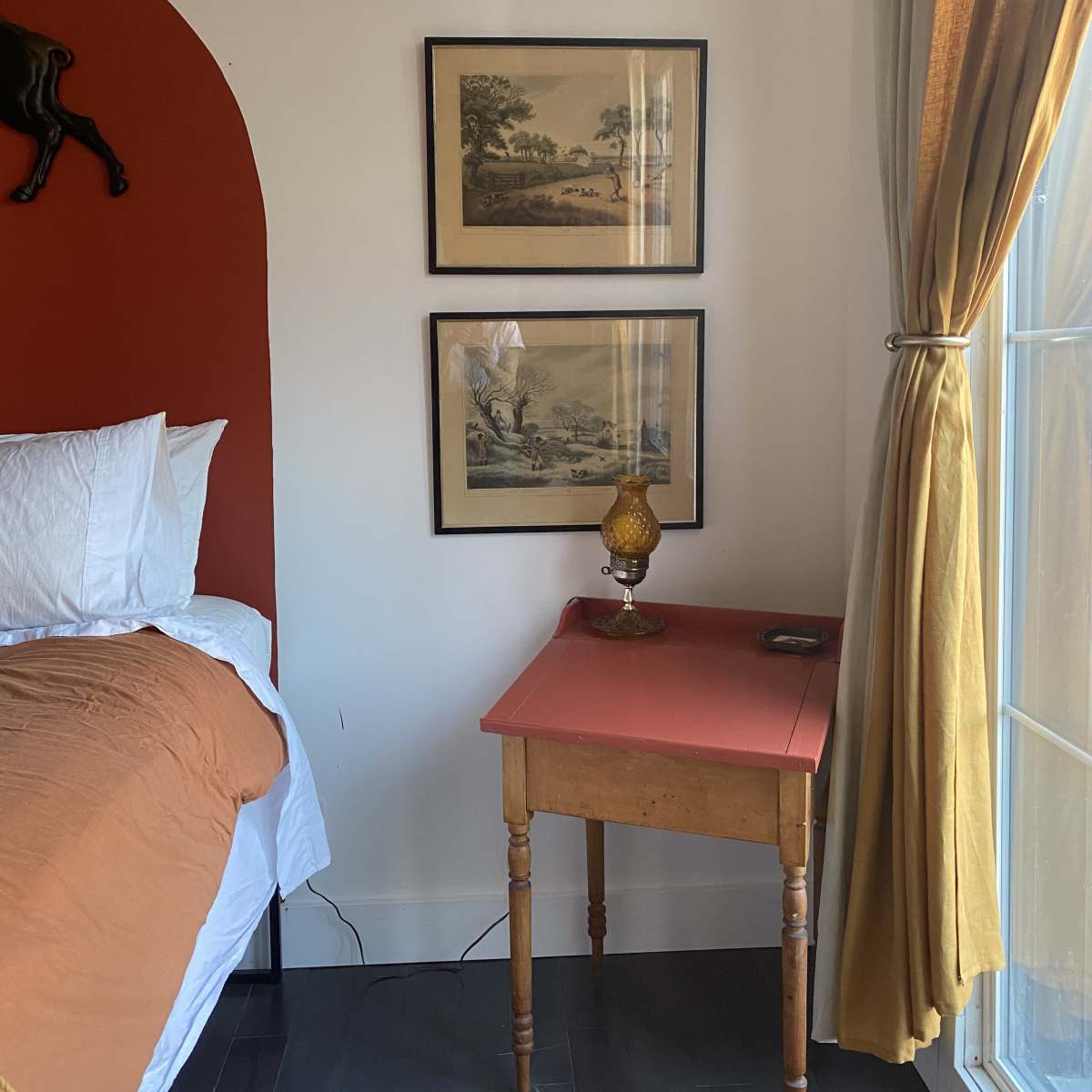 A bedroom corner featuring a bed with white linens, two framed pictures on the wall, a small table with a lamp, and a window with curtains.