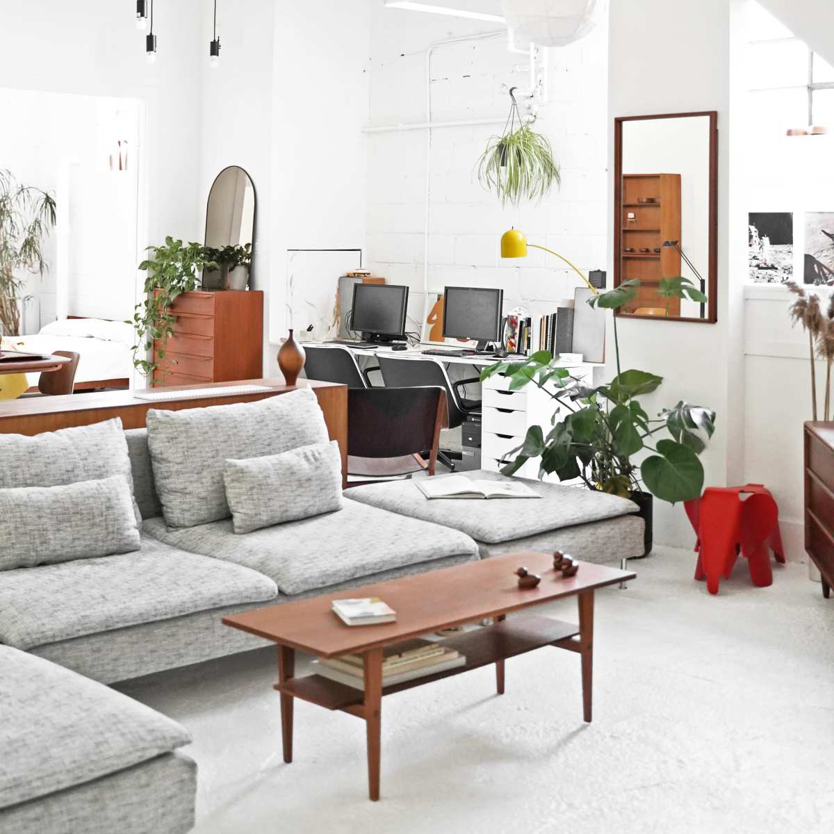 A modern living room features a sectional sofa, a wooden coffee table, plants, and a workspace with two computers in a bright, minimalist space.
