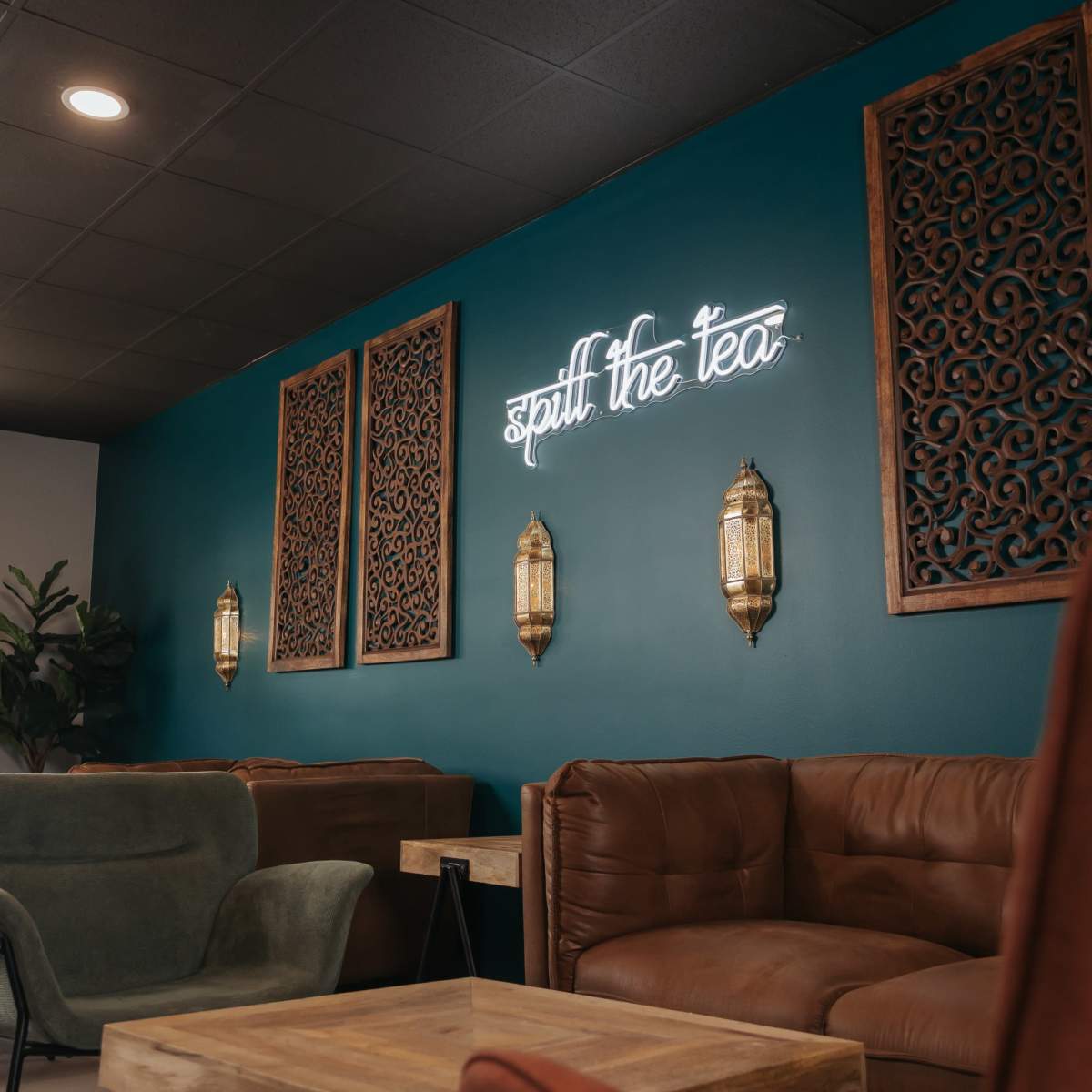 The interior of a cozy lounge features a teal wall with decorative panels and a neon sign that reads "Spill the Tea."