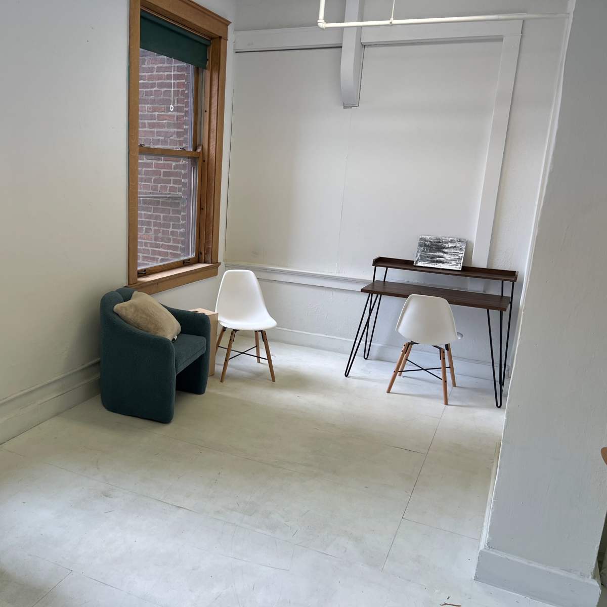 A room with a piano, a teal chair, a small desk, and two white chairs next to large windows.