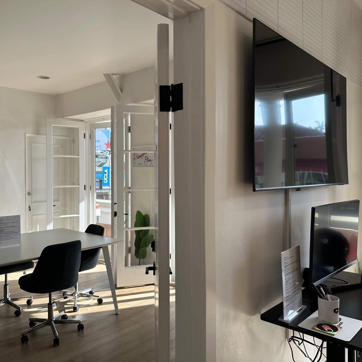 A modern office interior features a conference table, chairs, and a large screen, with sunlight streaming through open doors and windows.