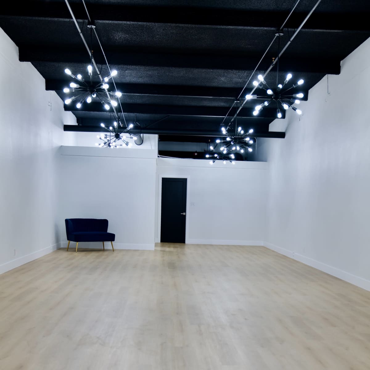 A spacious, empty room with light wooden flooring, white walls, and modern pendant lights, featuring a single dark blue chair in the corner.