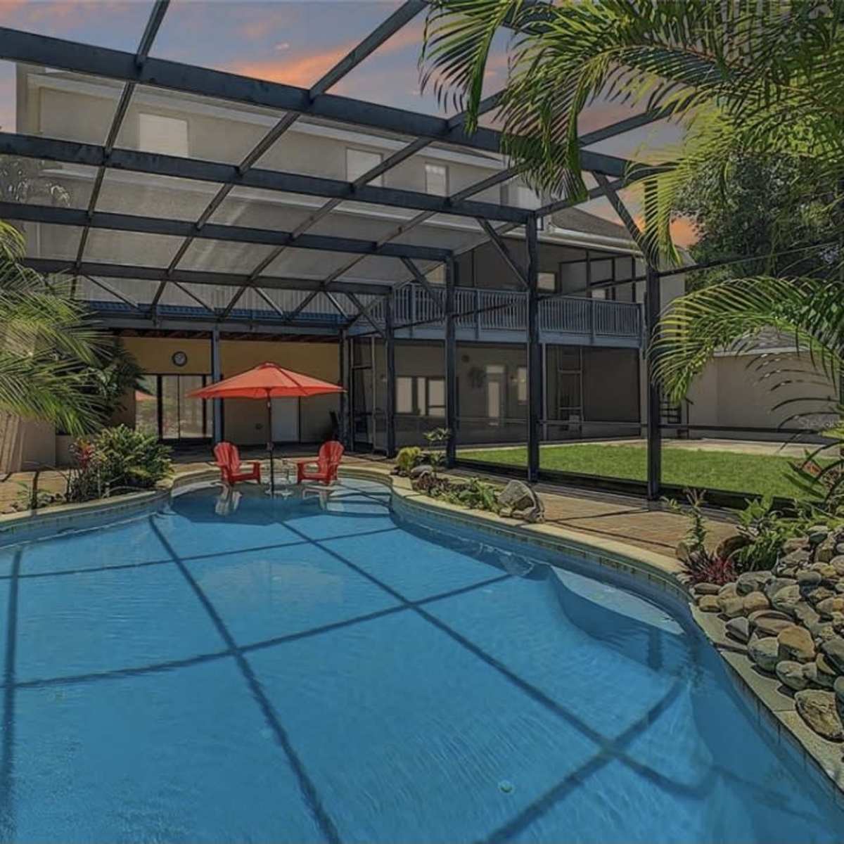 A swimming pool enclosed by a screened patio, featuring two red chairs and an umbrella beside the water.