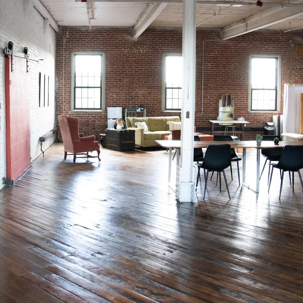 A spacious, empty room with wooden floors, large windows, and a dining area featuring a table and chairs, alongside a section with seating and cardboard boxes.