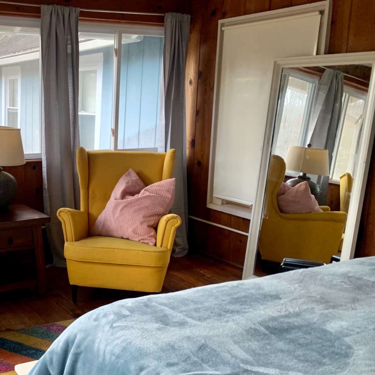 A cozy room with two yellow armchairs, a wooden nightstand with a lamp, and a large mirror, all surrounded by large windows.