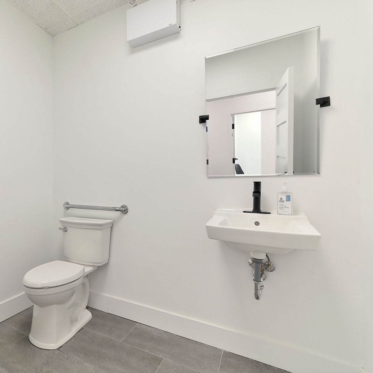A minimalistic bathroom featuring a toilet, a small sink, a mirror, and grab bars installed on the walls.
