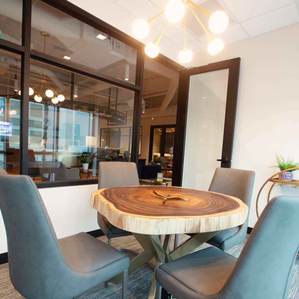 A round wooden table with four upholstered chairs sits in a modern conference room, illuminated by a contemporary light fixture.