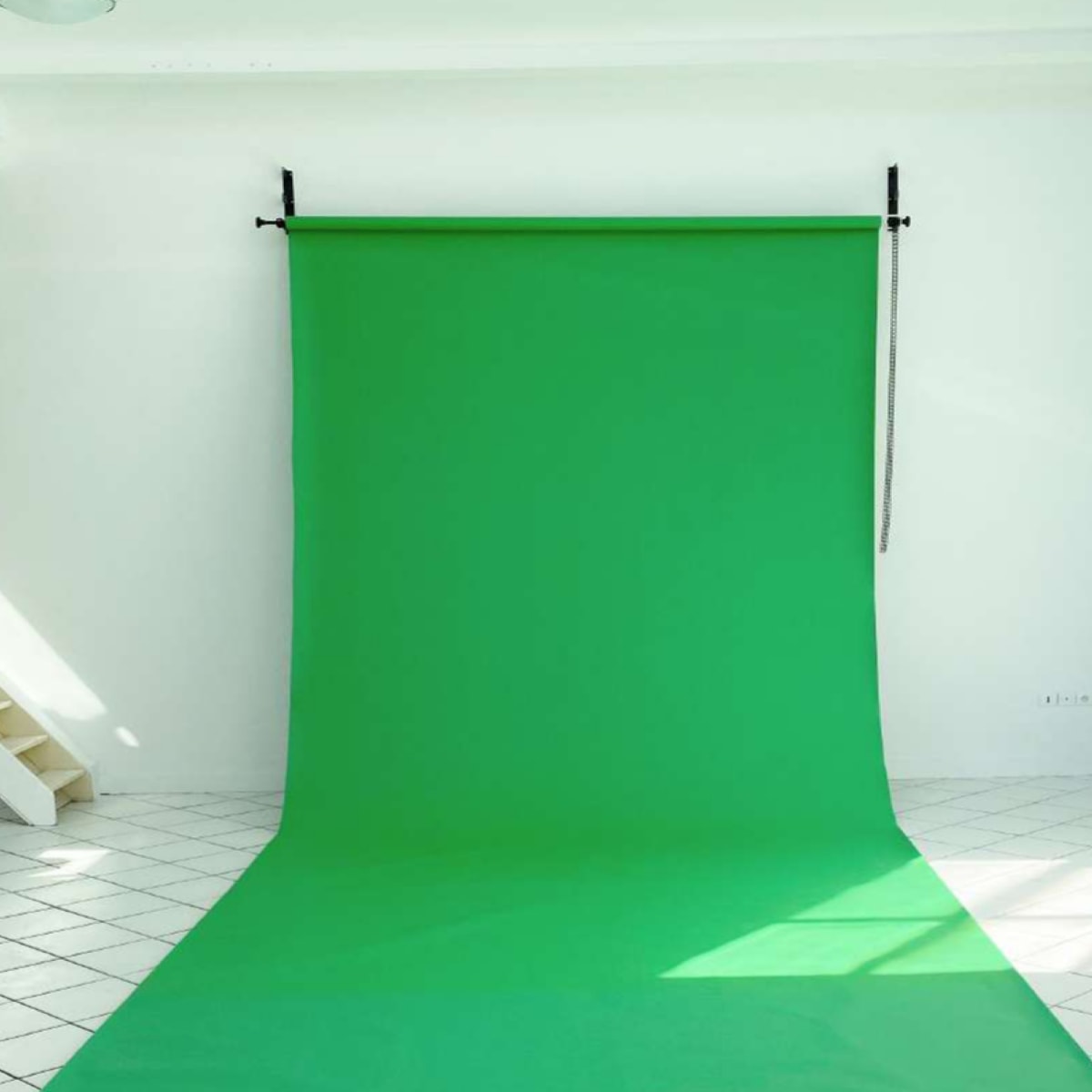 A green backdrop is set up in a brightly lit room with white walls and a staircase in the background.