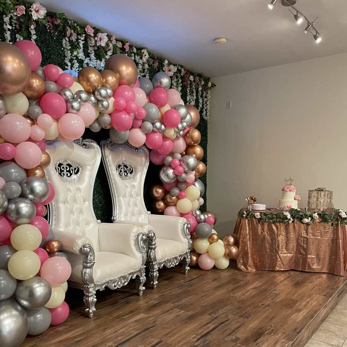 The scene features a decorated area with two white throne chairs surrounded by a colorful balloon arch and a table adorned with a pink tablecloth and cake.